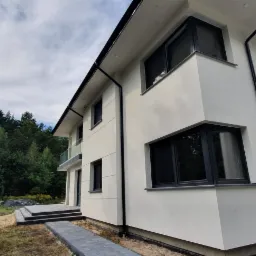 Dwukondygnacyjny dom z jasną elewacją, czarnymi ramami okiennymi i szarym brukowanym chodnikiem, otoczony zielenią drzew