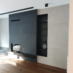 Nowoczesny salon z kominkiem obłożonym ciemnymi płytami i jasnymi płytami na ścianie, drewniana podłoga, czarne kwadratowe lampy sufitowe, czarne półki wnękowe z zegarem.