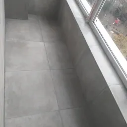 Szare płytki ceramiczne ułożone na podłodze i ścianach balkonu, widok z góry na narożnik przy oknie.