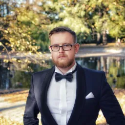 Mężczyzna w muszce i garniturze na tle jesiennego parku z jeziorkiem. Elegancki strój, okulary i broda. Portret w plenerze, ciepłe barwy jesieni.