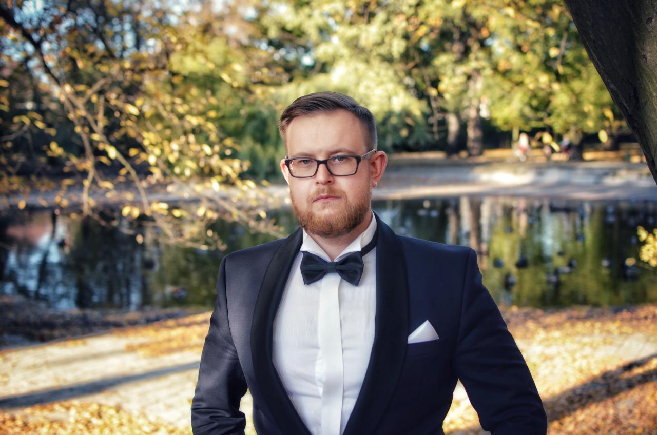 Mężczyzna w muszce i garniturze na tle jesiennego parku z jeziorkiem. Elegancki strój, okulary i broda. Portret w plenerze, ciepłe barwy jesieni.