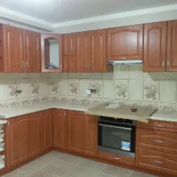 Kompletna zabudowa kuchenna z drewnianymi szafkami, płytkami ceramicznymi z motywem filiżanek i owoców oraz piekarnikiem zabezpieczonym kartonem.