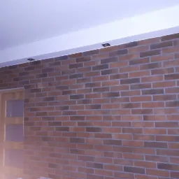 Ściana z cegieł klinkierowych w odcieniach brązu i szarości, z zamontowanymi punktowymi lampami sufitowymi i częściowo widocznymi drewnianymi drzwiami z trzema szybami.