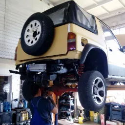 Żółty samochód terenowy, podniesiony na podnośniku, z widocznym pracownikiem w kombinezonie, naprawiającym tylny most. Czerwone sprężyny zawieszenia i koło zapasowe z tyłu.