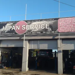 Fasada warsztatu samochodowego Japan Service z trzema stanowiskami, na dwóch podnośnikach auta w trakcie naprawy, na elewacji baner reklamowy z zakresem usług i numerami telefonów.
