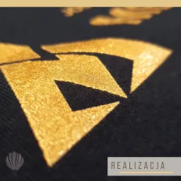Zbliżenie na złoty, geometryczny wzór na czarnym materiale, prawdopodobnie koszulce, z efektem połysku i cieniowania, sugerujące wysoką jakość wykonania nadruku.