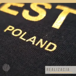 Czarny materiał z bliska, z naniesionym złotym nadrukiem 'EST POLAND', wykonany techniką flock.