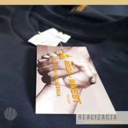 Ciemna koszulka z metką Gildan i przywieszką z logo Mega-Invest Poland oraz hasłem 'Ludzie Godni Polecenia', widoczny napis 'Realizacja'.