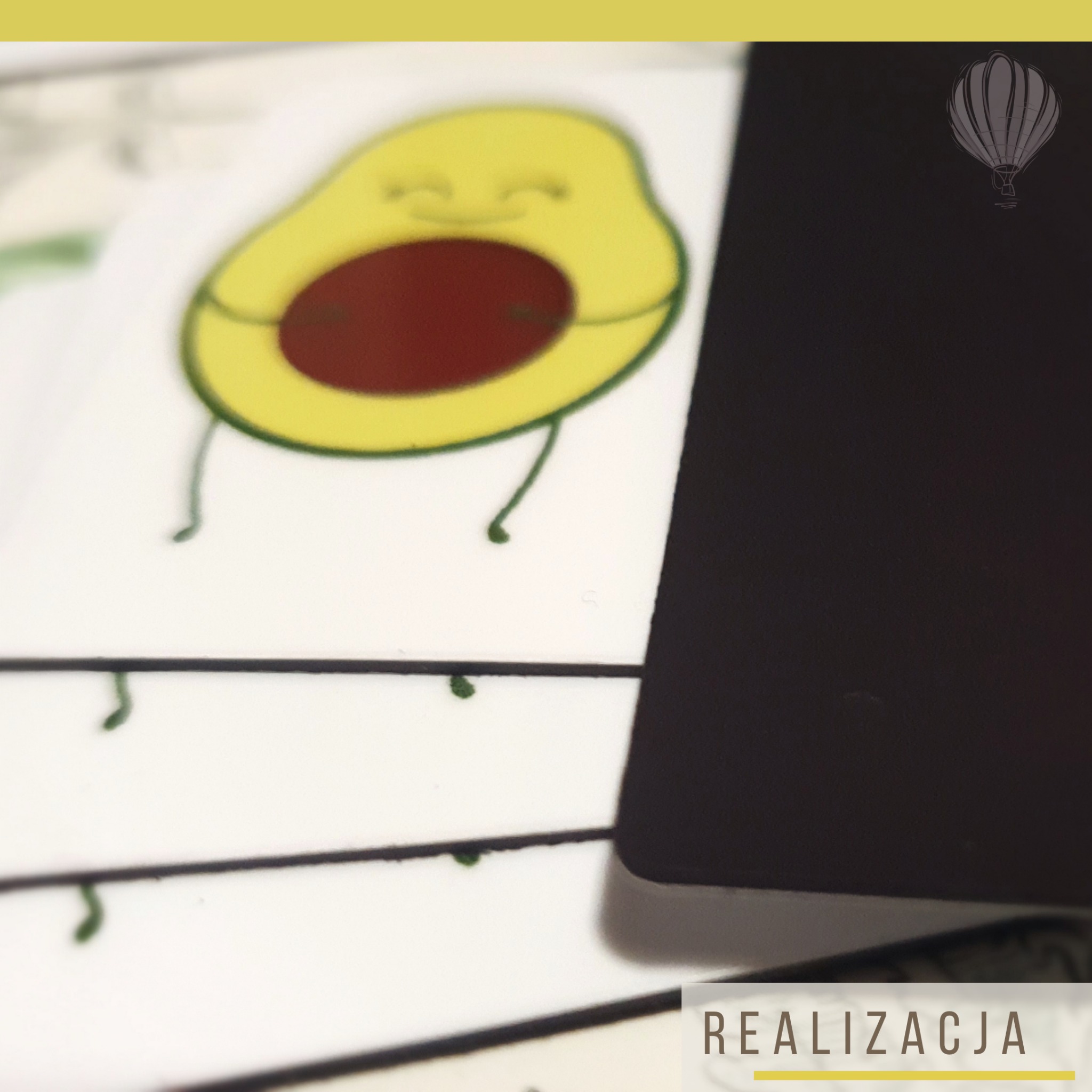 Stos magnesów z grafiką awokado z rączkami i uśmiechniętą buzią, jeden magnes częściowo zasłonięty czarnym magnesem z szarą grafiką balonu na ogrzane powietrze w prawym górnym rogu.