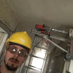 Pracownik w kasku i okularach ochronnych, wykonujący odwiert w ścianie za pomocą wiertnicy diamentowej, widoczna drabina i przewody elektryczne.