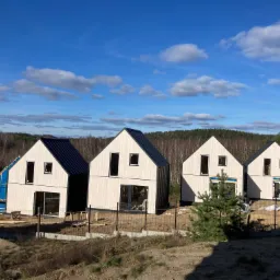 Cztery nowoczesne domy w trakcie budowy, z drewnianą elewacją i czarnym dachem, otoczone ogrodzeniem z siatki, na tle lasu i błękitnego nieba z chmurami.