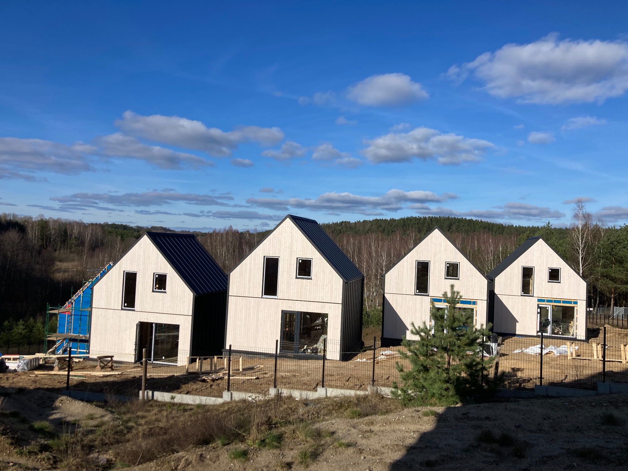Cztery nowoczesne domy w trakcie budowy, z drewnianą elewacją i czarnym dachem, otoczone ogrodzeniem z siatki, na tle lasu i błękitnego nieba z chmurami.