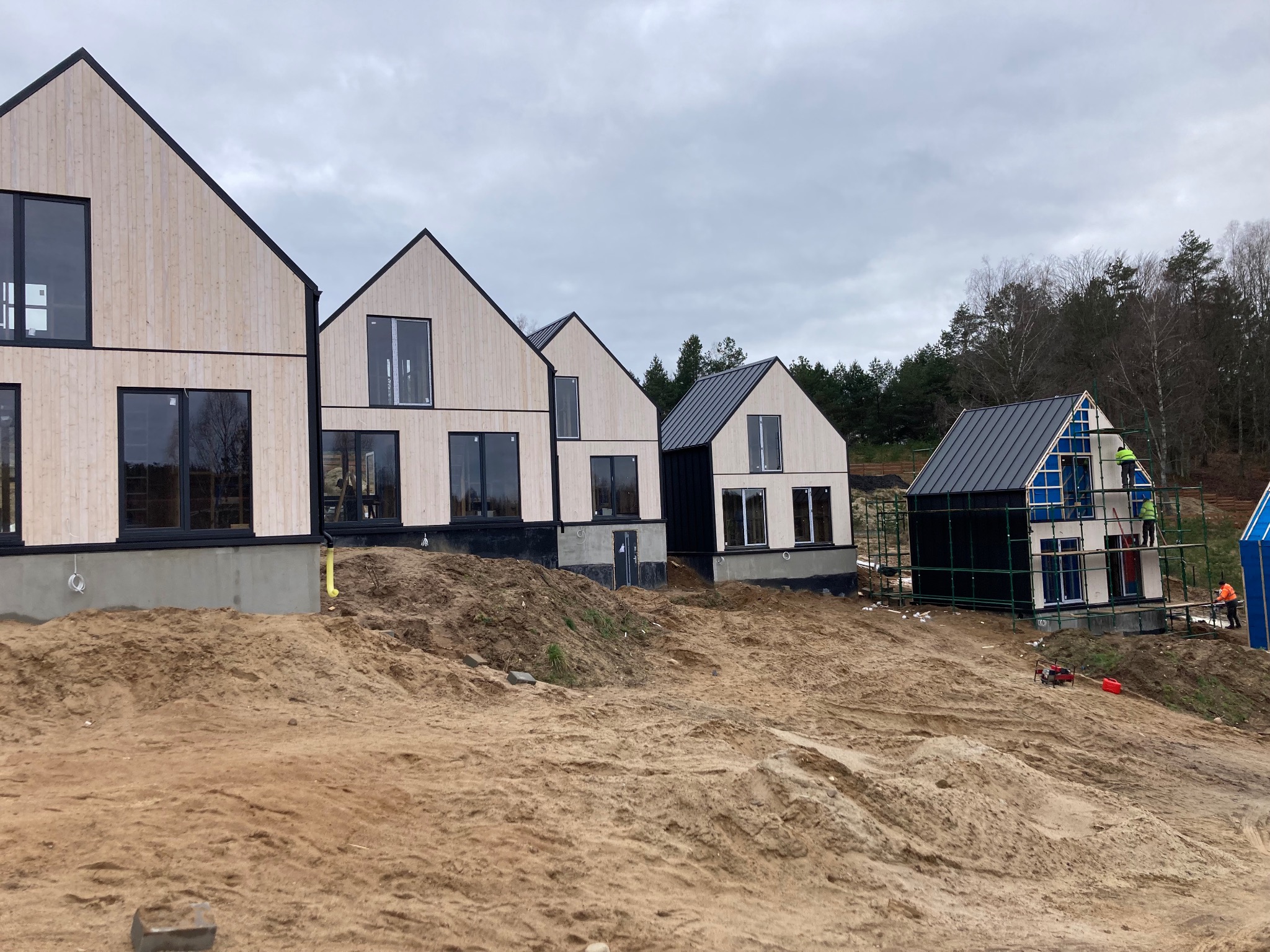 Nowoczesne domy w stylu skandynawskim z jasną elewacją drewnianą i czarnymi ramami okiennymi, w trakcie budowy, z widocznymi rusztowaniami i pracownikami na tle lasu i pochmurnego nieba.