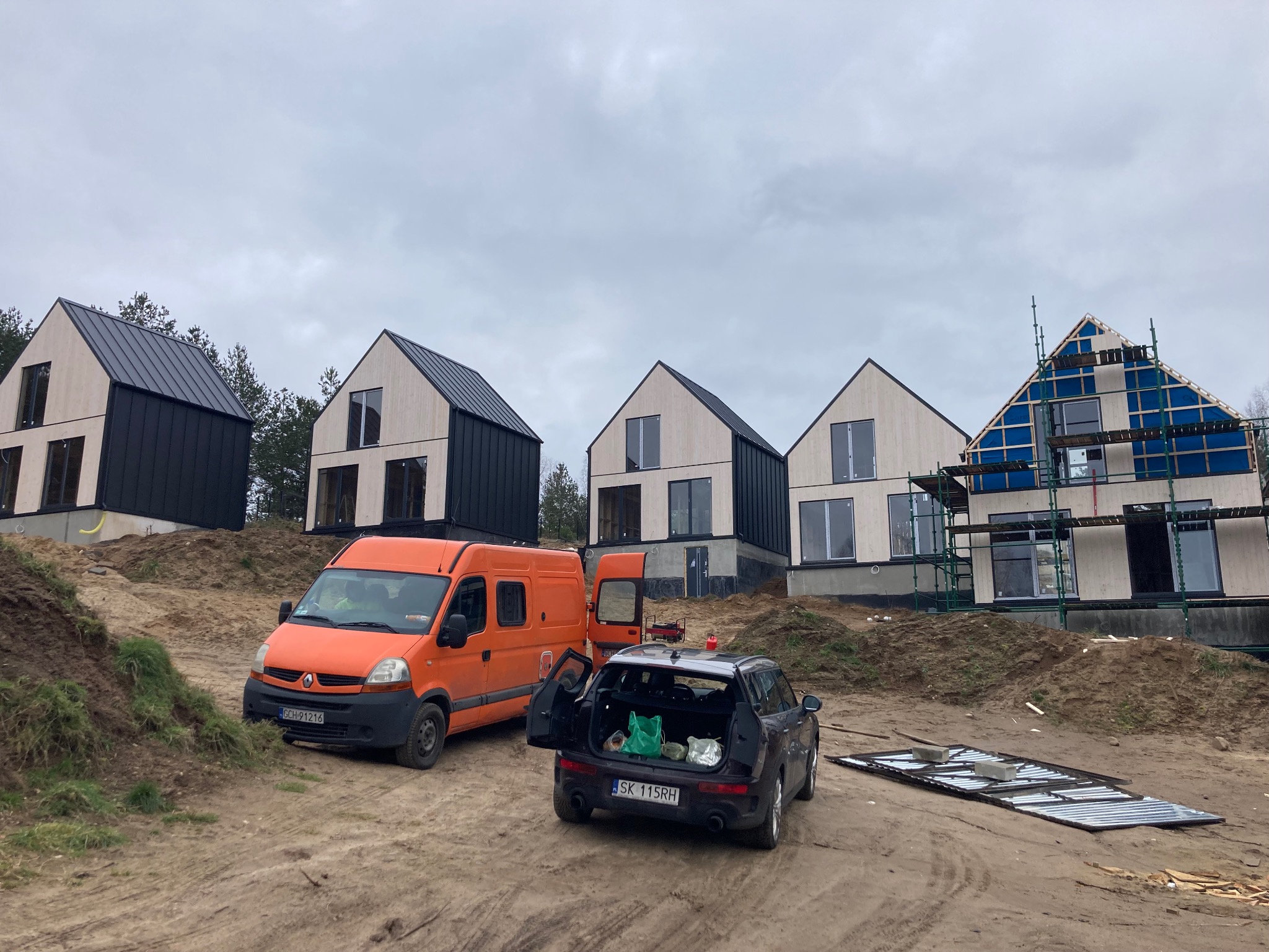 Nowoczesne domy w trakcie budowy, z pomarańczowym busem i czarnym samochodem osobowym z otwartym bagażnikiem na pierwszym planie.