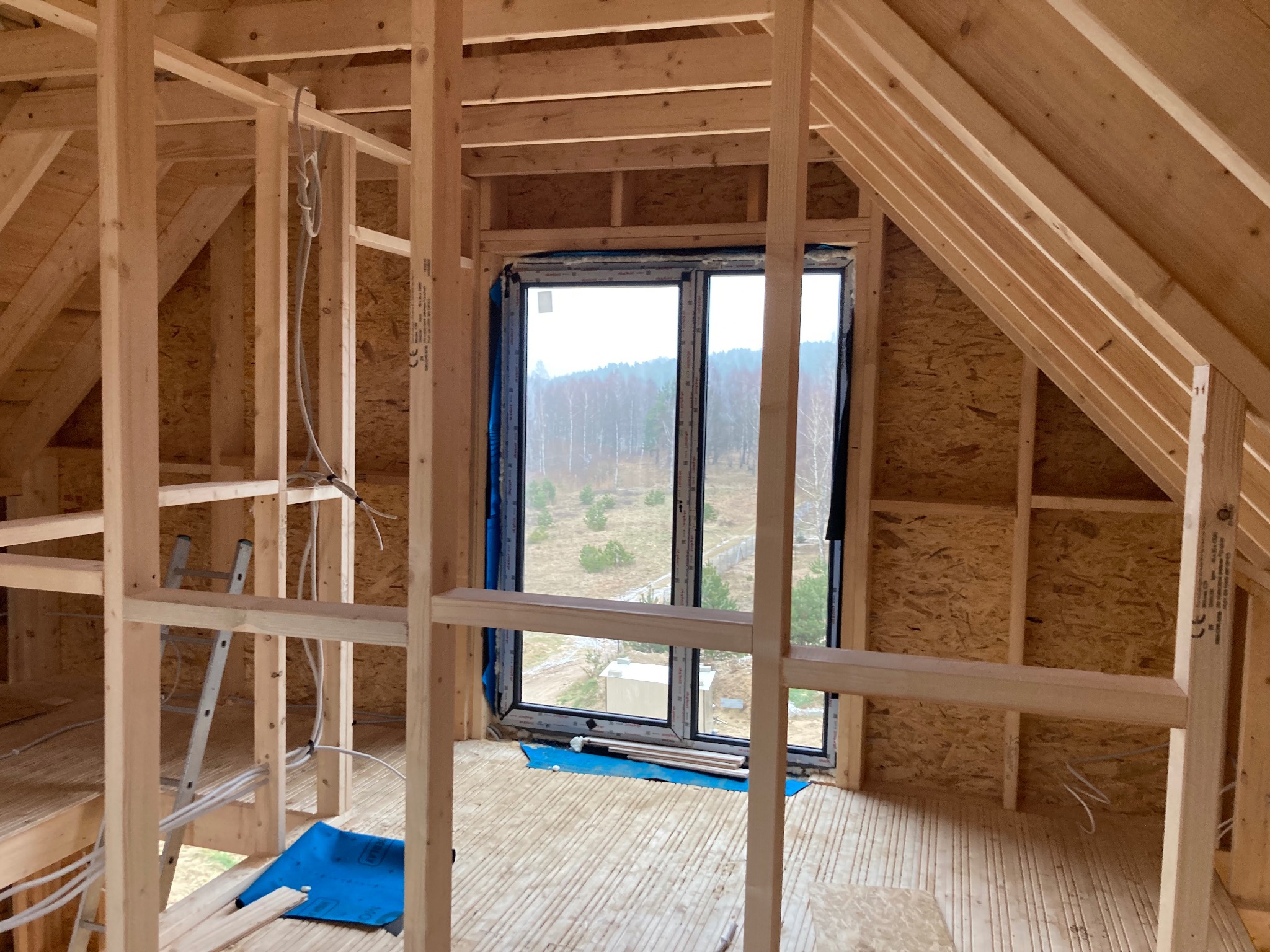 Wnętrze poddasza w trakcie budowy z widoczną drewnianą konstrukcją szkieletową, oknem z widokiem na zewnątrz i częściowo ułożoną podłogą z desek.