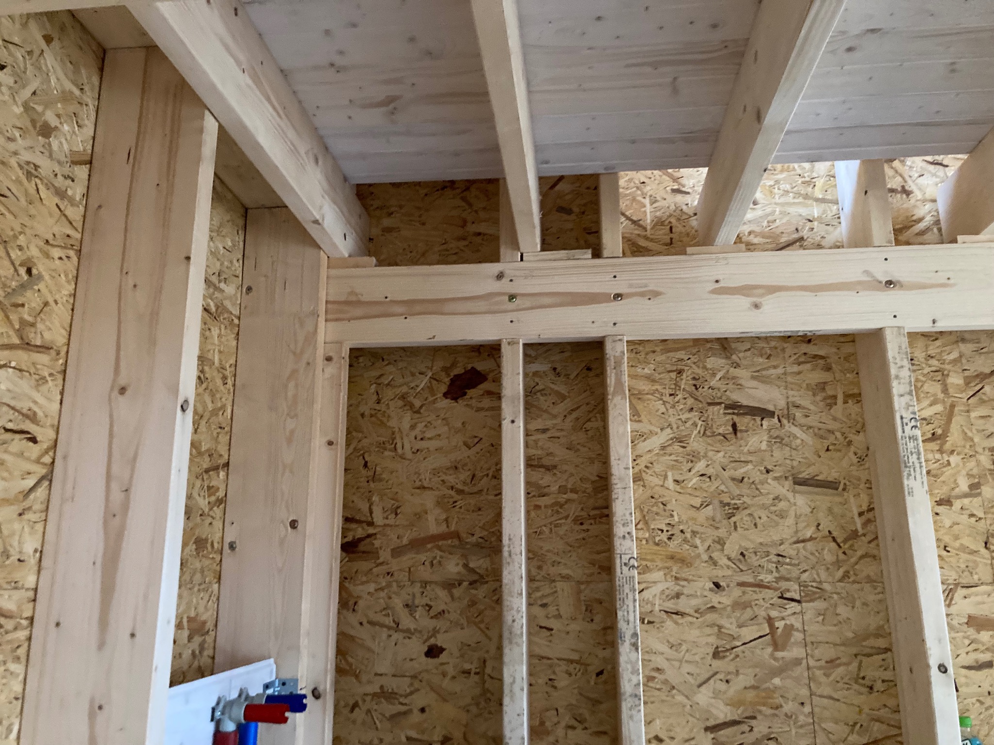 Konstrukcja szkieletowa ścian z płyt OSB, widoczne drewniane belki stropowe i instalacja wodna z rurami czerwonymi i niebieskimi.