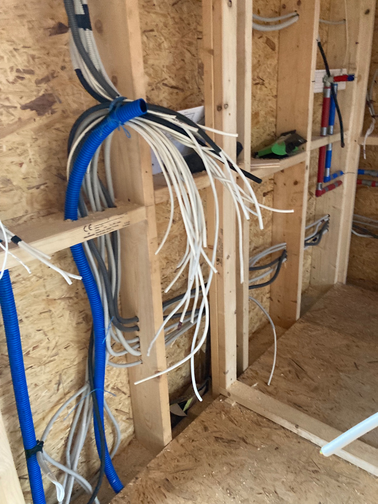 Wiązka niebieskich, szarych i białych kabli oraz rur, częściowo poprowadzonych w drewnianej konstrukcji ściany w surowym stanie.