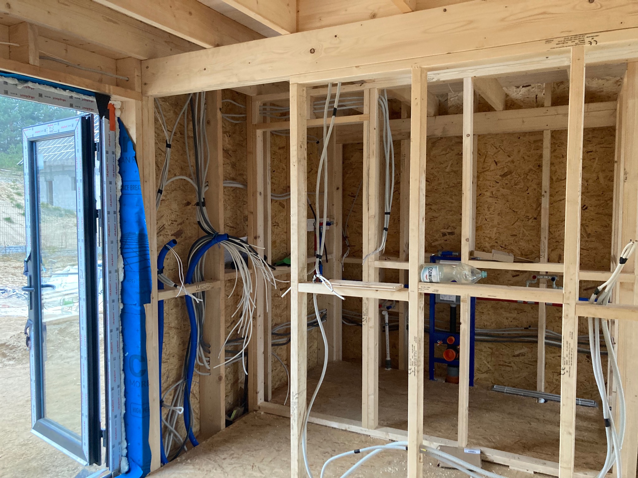 Wnętrze domu w trakcie budowy: drewniana konstrukcja szkieletowa, instalacje elektryczne i hydrauliczne w trakcie montażu, widoczne okno z niebieską folią ochronną oraz butelka wody na konstrukcji.