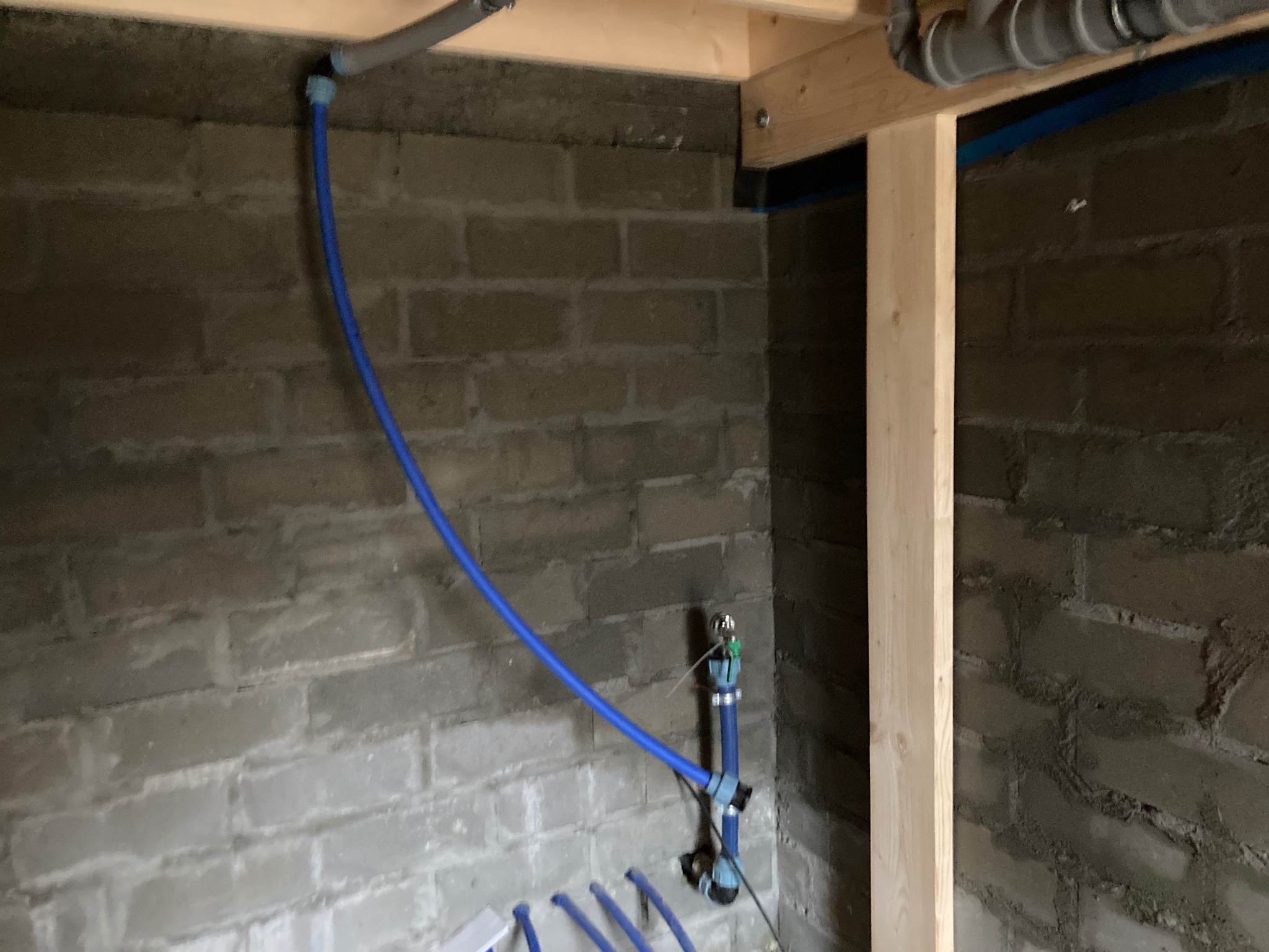 Fragment surowej ściany z szarej cegły z niebieskimi rurami instalacji wodnej oraz drewnianą konstrukcją wsporczą.