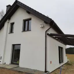 Nowoczesny, biały dom z ciemnobrązowym dachem i stolarką okienną, widok z boku na świeżo otynkowaną elewację z elementami instalacji na zewnątrz, w tle pola i las.