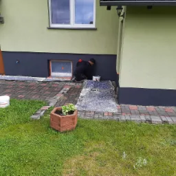 Mężczyzna w czarnym ubraniu maluje dolną część elewacji domu na ciemny kolor, zabezpieczone okno w tle, wiadro z farbą obok, trawnik z kostką brukową z przodu.