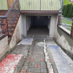 Przygotowanie do izolacji fundamentów: odkopane fundamenty, częściowo oczyszczone, z widocznymi śladami wilgoci i fragmentami starej izolacji, przykryte folią ochronną, z kostką brukową częściowo...