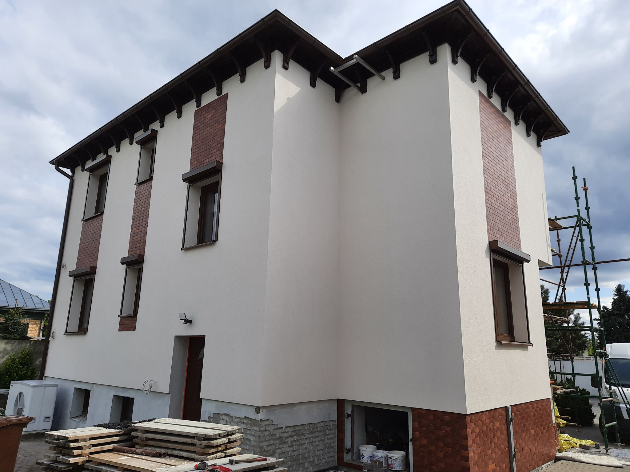 Dwupiętrowy dom z jasną elewacją i ceglanymi wstawkami, widoczne rusztowania i materiały budowlane, częściowo wykończony cokół.