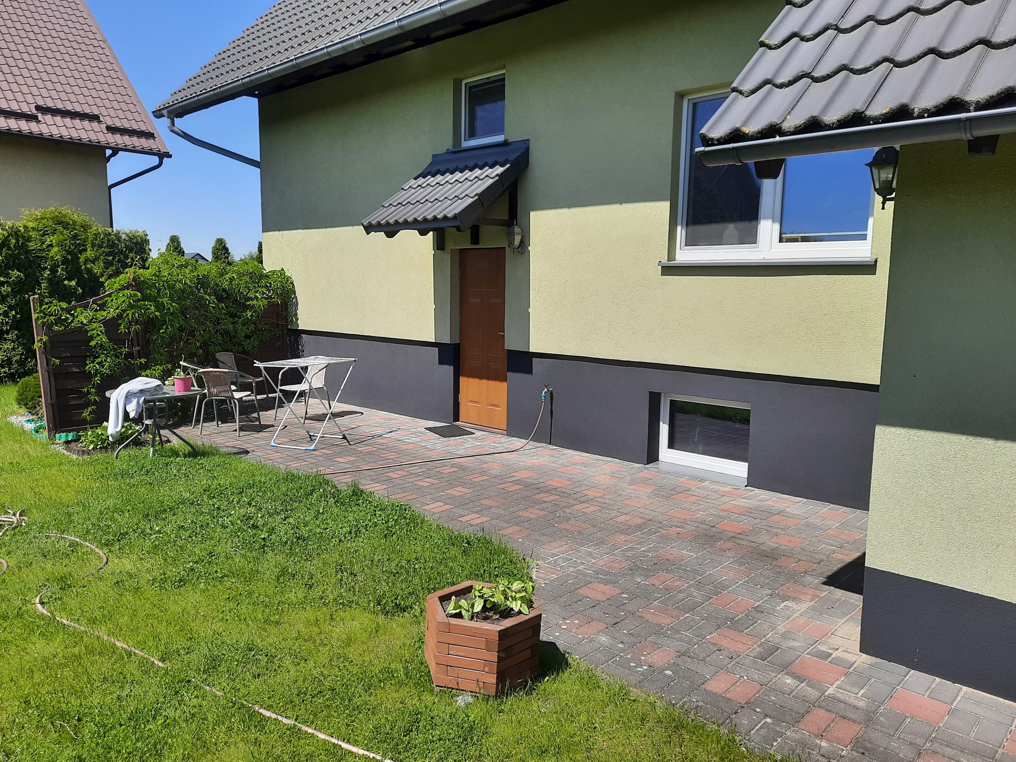 Ujęcie domu jednorodzinnego z ogrodem: widoczna elewacja w odcieniach zieleni i szarości, kostka brukowa, trawnik z wężem ogrodowym, meble ogrodowe i donica z roślinami. Elewacja po renowacji.