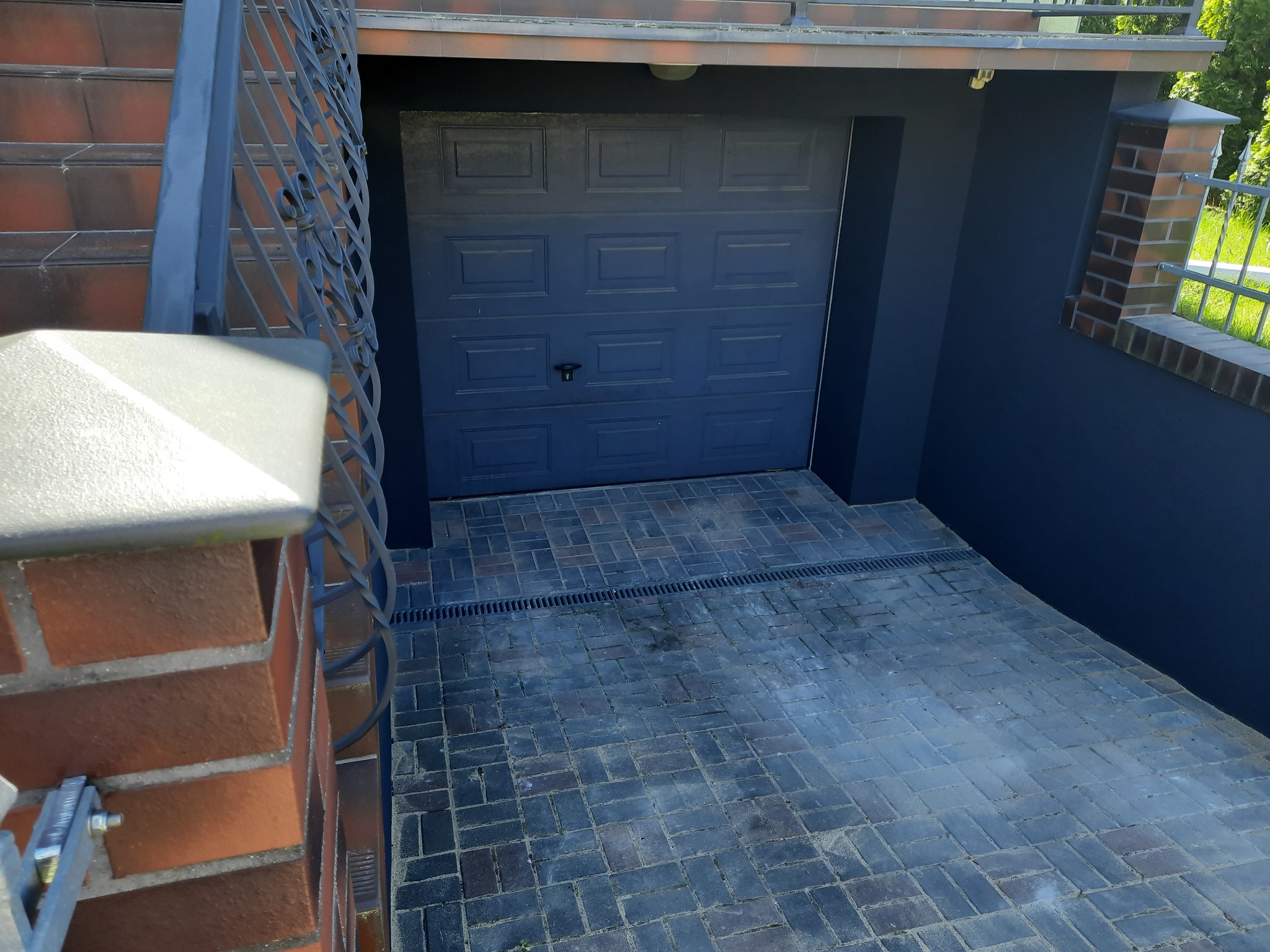 Brama garażowa w kolorze grafitowym, elewacja w odcieniach szarości, podjazd wyłożony kostką brukową z odpływem liniowym, widoczne fragmenty schodów z cegły i metalowej balustrady.