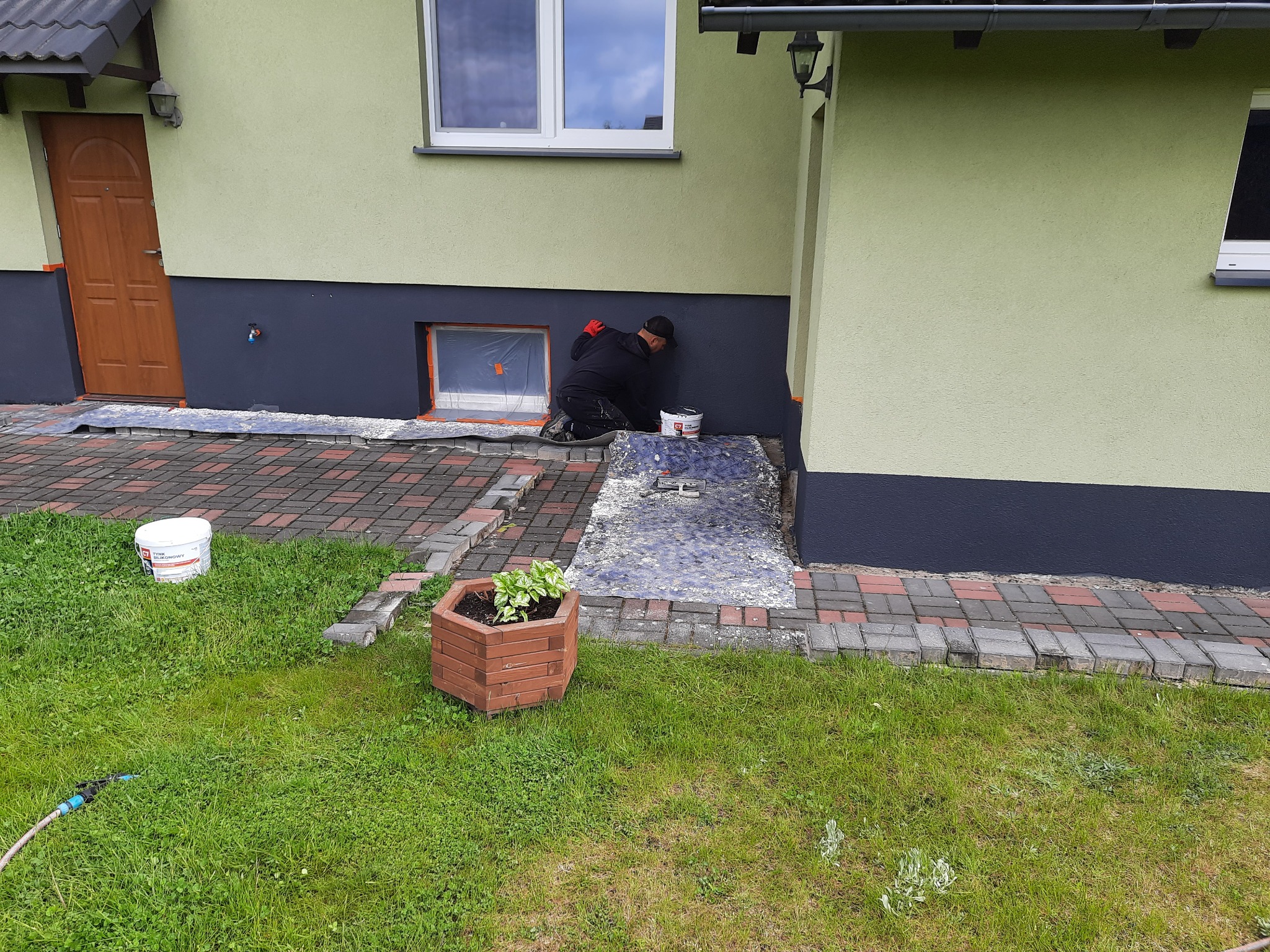 Mężczyzna w czarnym ubraniu maluje dolną część elewacji domu na ciemny kolor, zabezpieczone okno w tle, wiadro z farbą obok, trawnik z kostką brukową z przodu.