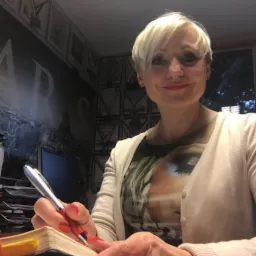 Kobieta z krótkimi blond włosami pisze w notatniku srebrnym długopisem, w tle ściana z motywem Paryża.