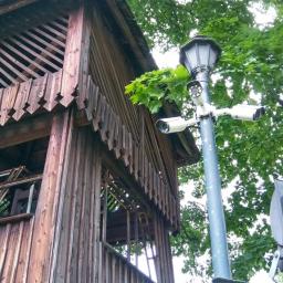 FHU Sylen Antoni Strózik - Trzy kamery monitoringu zamontowane na słupie oświetleniowym, obok drewnianej konstrukcji w parku, widoczne liście drzew.