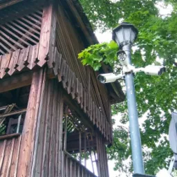 Trzy kamery monitoringu zamontowane na słupie oświetleniowym, obok drewnianej konstrukcji w parku, widoczne liście drzew.