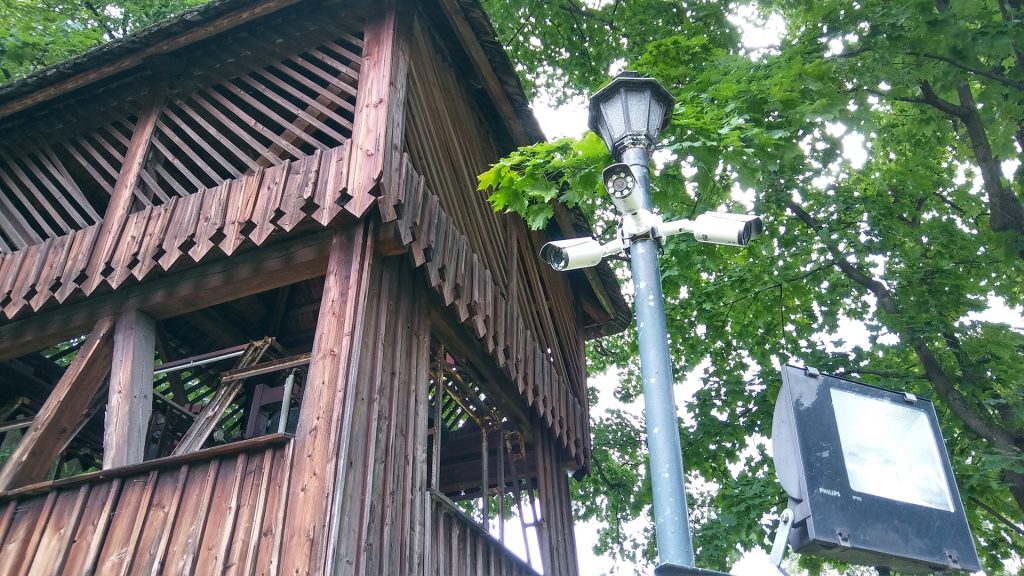 Trzy kamery monitoringu zamontowane na słupie oświetleniowym, obok drewnianej konstrukcji w parku, widoczne liście drzew.