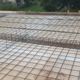 Zbrojenie stropu na deskowaniu z widocznymi prętami pionowymi i poziomymi, przygotowane do zalania betonem, w tle zieleń.