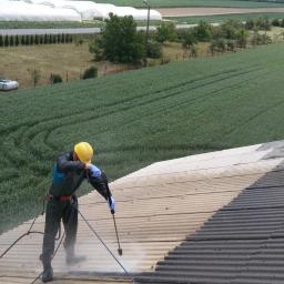 Wash-King Malowanie dachów Łukasz Biernat - Pracownik w kasku i kombinezonie czyści dach z eternitu za pomocą myjki ciśnieniowej, widoczne różnice w kolorze między czystą a brudną powierzchnią dachu, w tle pole uprawne i szklarnie.