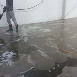 Wash-King Malowanie dachów Łukasz Biernat - Pracownik w gumowych butach czyści posadzkę przemysłową maszyną z czerwonym elementem, widoczne ślady detergentu i mokra powierzchnia.