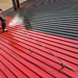 Pracownik w kombinezonie zabezpieczającym, z linami i uprzężą, czyści ciśnieniowo dach z blachy trapezowej, którego część jest już pomalowana na szaro, a część czeka na malowanie i ma kolor czerwony.