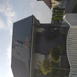 Pracownik w kasku, zabezpieczony linami, zjeżdża po stromej, szarej połaci dachu domu jednorodzinnego w słoneczny dzień, widok z dołu.