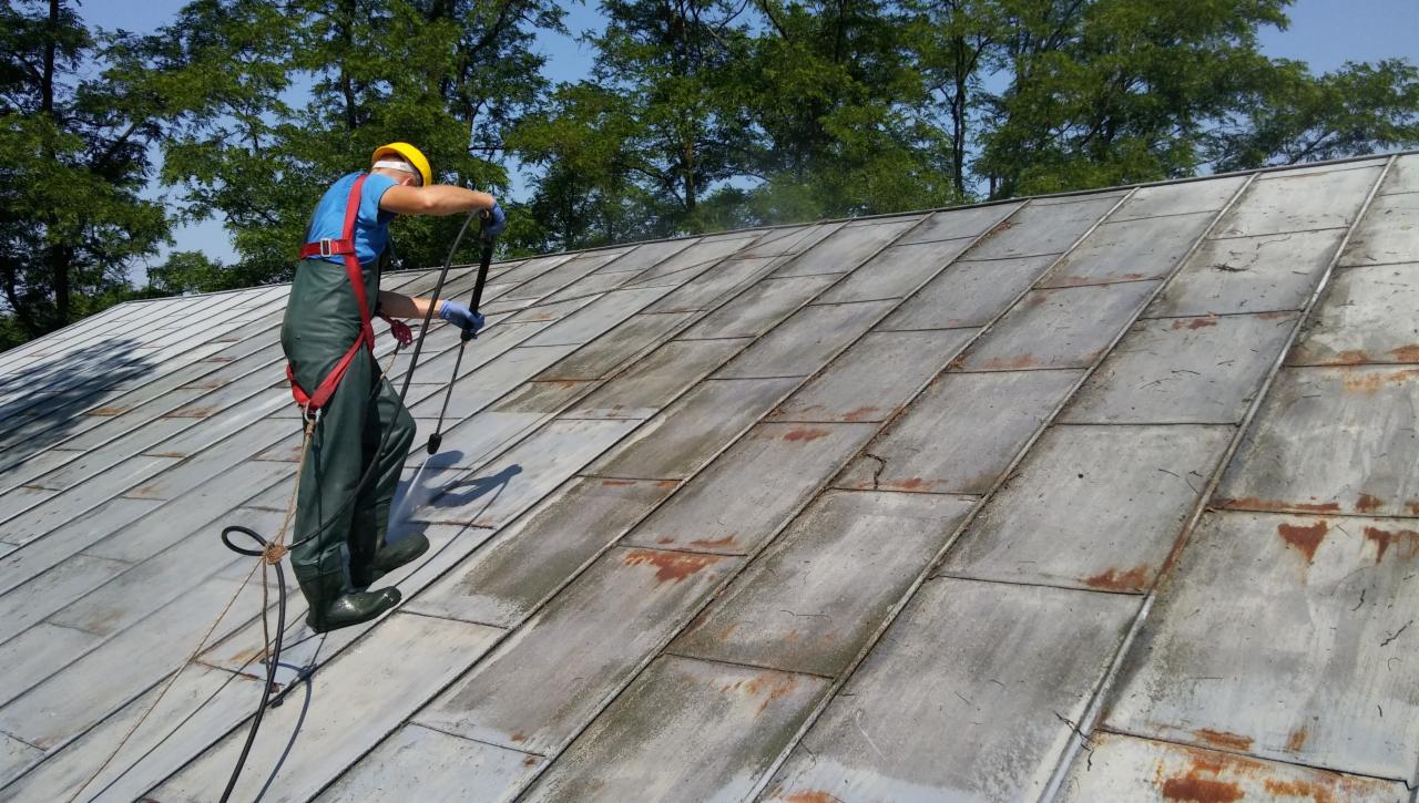 Pracownik w kombinezonie i kasku zabezpieczony linami czyści metalowy dach z rdzą za pomocą myjki ciśnieniowej, widoczne drzewa w tle.