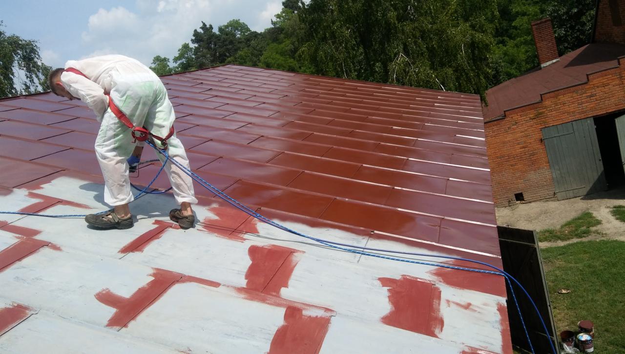Pracownik w kombinezonie maluje dach z blachy na brązowo za pomocą pistoletu natryskowego, widoczne fragmenty dachu pokryte podkładem, w tle budynek gospodarczy z cegły.