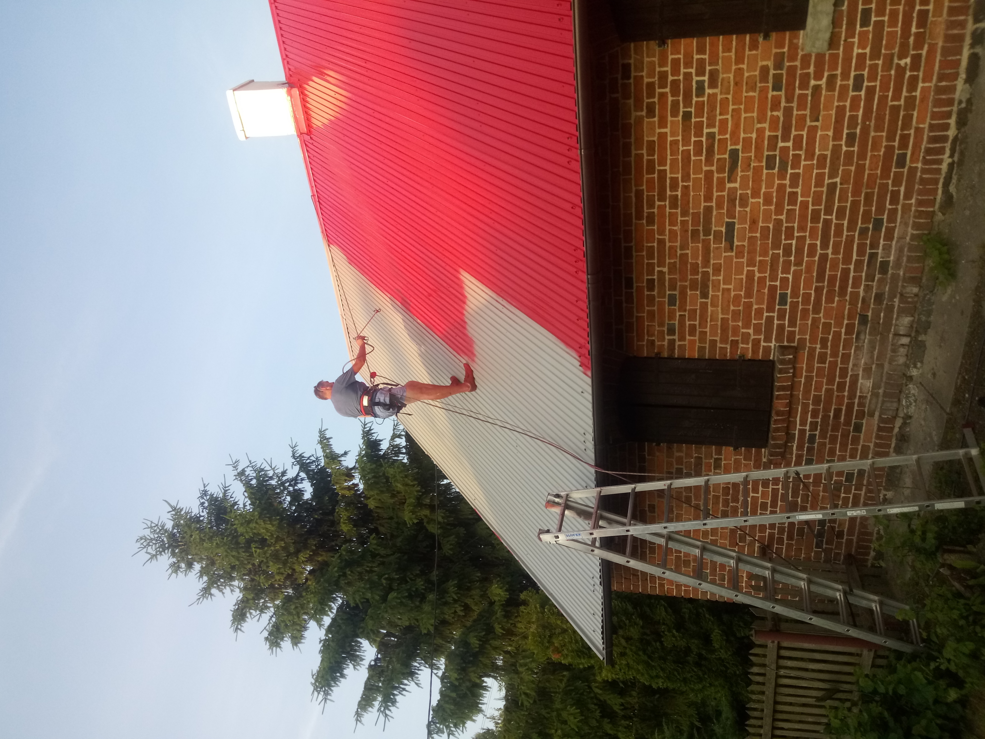 Malowanie dachu z blachy trapezowej na czerwono, widoczny pracownik zabezpieczony linami, drabina oparta o ceglaną ścianę budynku, częściowo pomalowany dach.