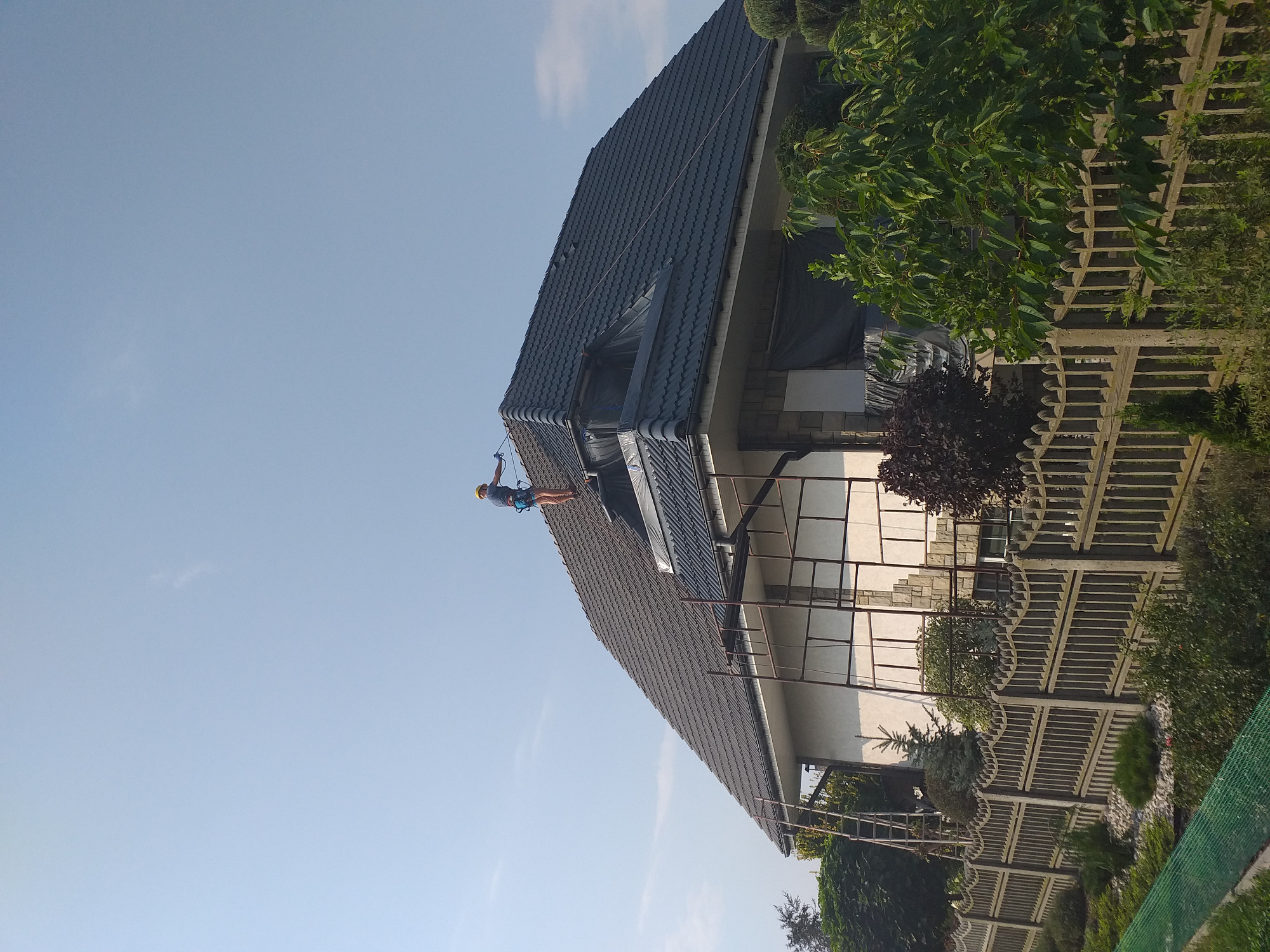 Pracownik w kasku, zabezpieczony linami, wykonuje prace na wysokości na skośnym dachu z ciemnej dachówki, widoczne rusztowanie i fragment ogrodu z ozdobnym płotem.