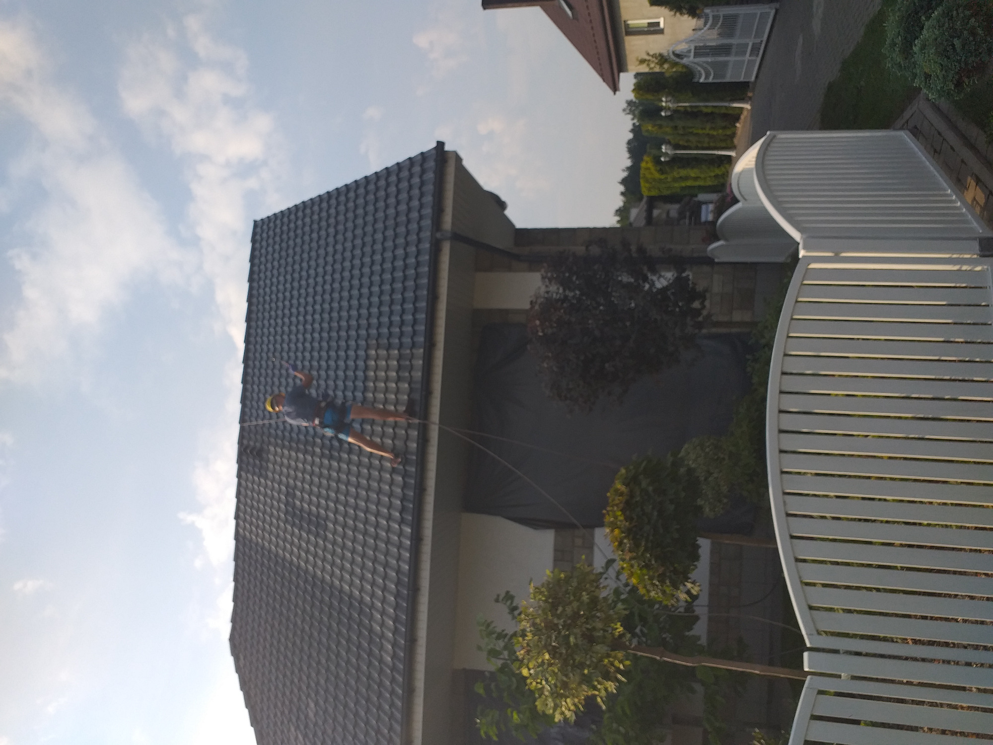 Pracownik zabezpieczony linami na spadzistym dachu krytym ciemną dachówką, widok z dołu, fragment białego ogrodzenia w kadrze.