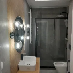 Nowoczesna łazienka z umywalką nablatową, owalnym lustrem z oświetleniem kulowym, kabiną prysznicową ze szklanymi drzwiami i podświetlanym sufitem.