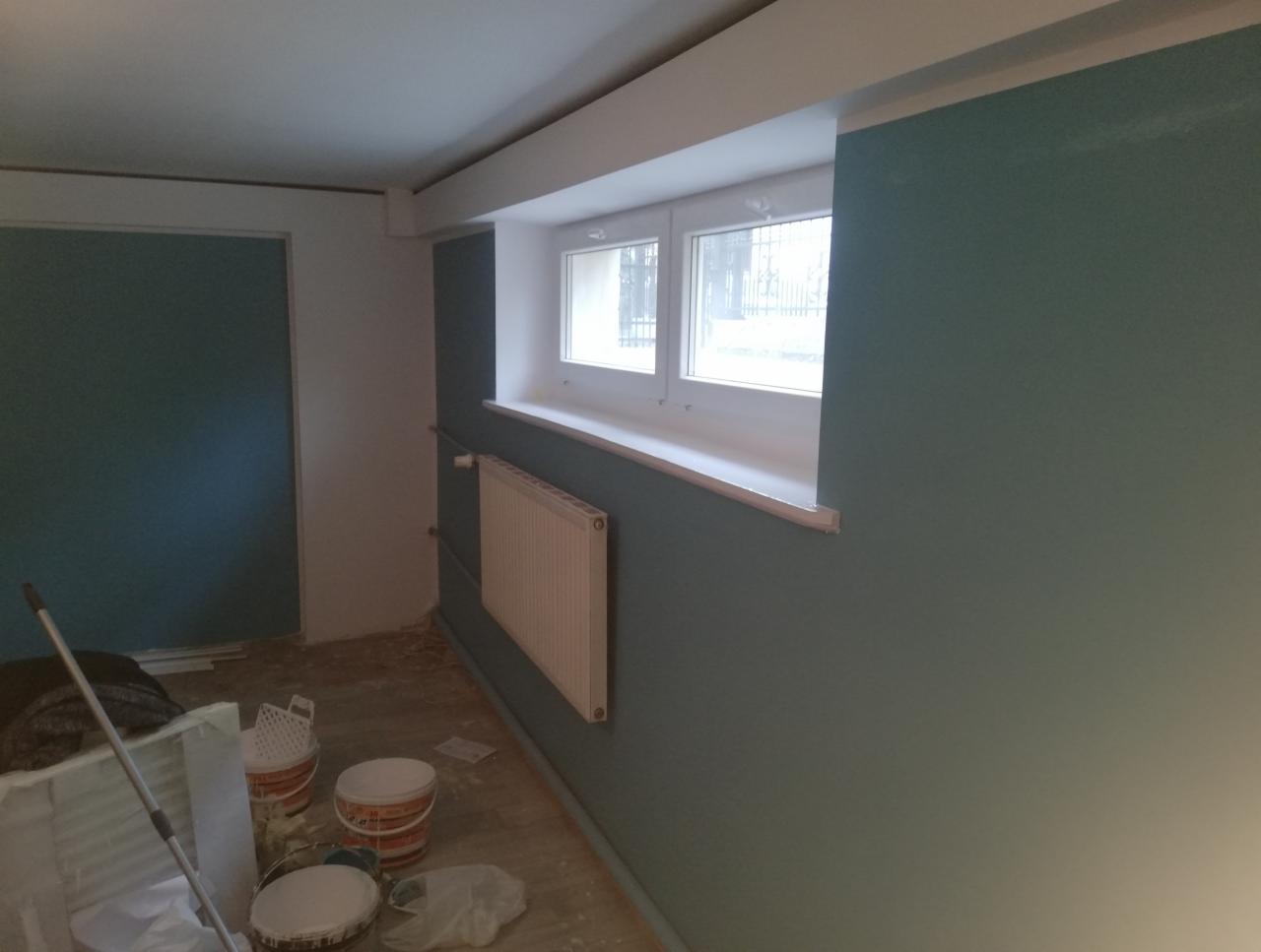 Wnętrze w trakcie malowania na odcień zieleni, widoczne puszki z farbą, grzejnik pod oknem i częściowo pomalowane ściany.