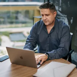 Mężczyzna w niebieskiej koszuli skupiony na pracy na laptopie Apple, obok telefon i dokumenty na okrągłym drewnianym stole, z widokiem na zieleń za oknem i artystyczną grafiką na ścianie w tle.