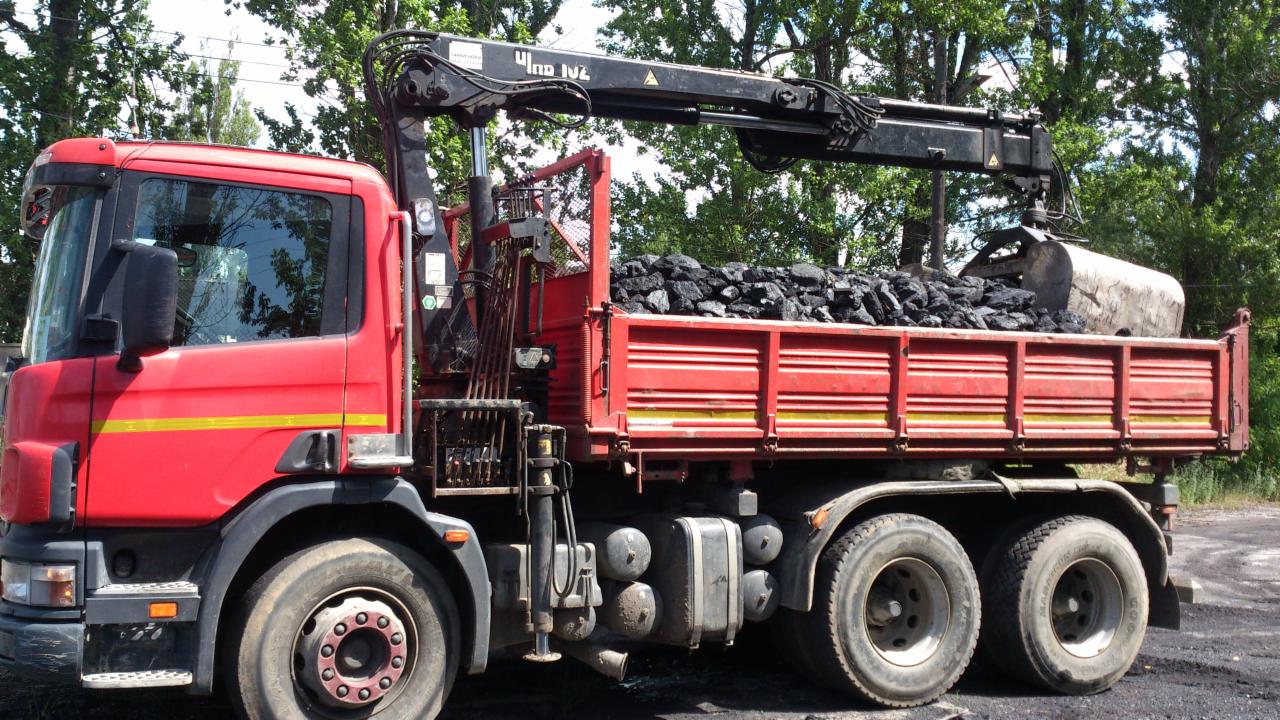 Czerwona ciężarówka z hydraulicznym żurawiem załadowana węglem, widok z boku na tle zieleni drzew.