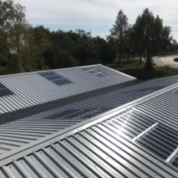 Dwa budynki z szarym dachem z blachy trapezowej, w dachu wbudowane okna dachowe i panele słoneczne, w tle drzewa i droga z samochodami.