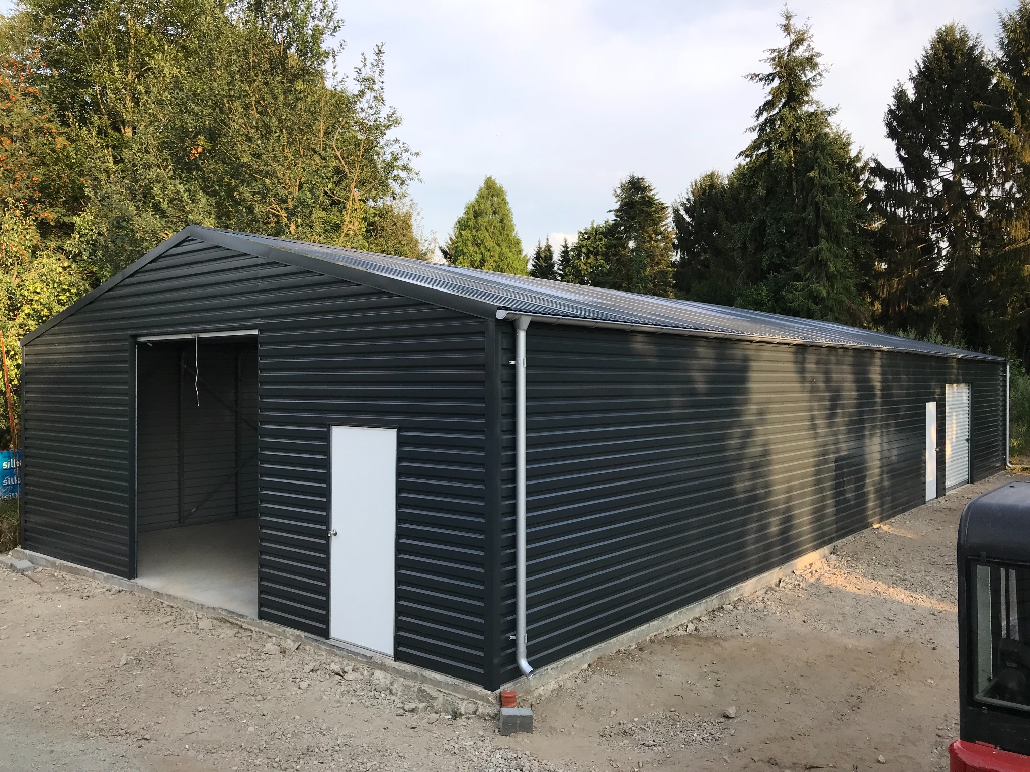 Nowoczesna, długa hala z czarnej blachy trapezowej z białymi drzwiami, otwartymi wrotami i betonową wylewką, otoczona drzewami i gruntem.